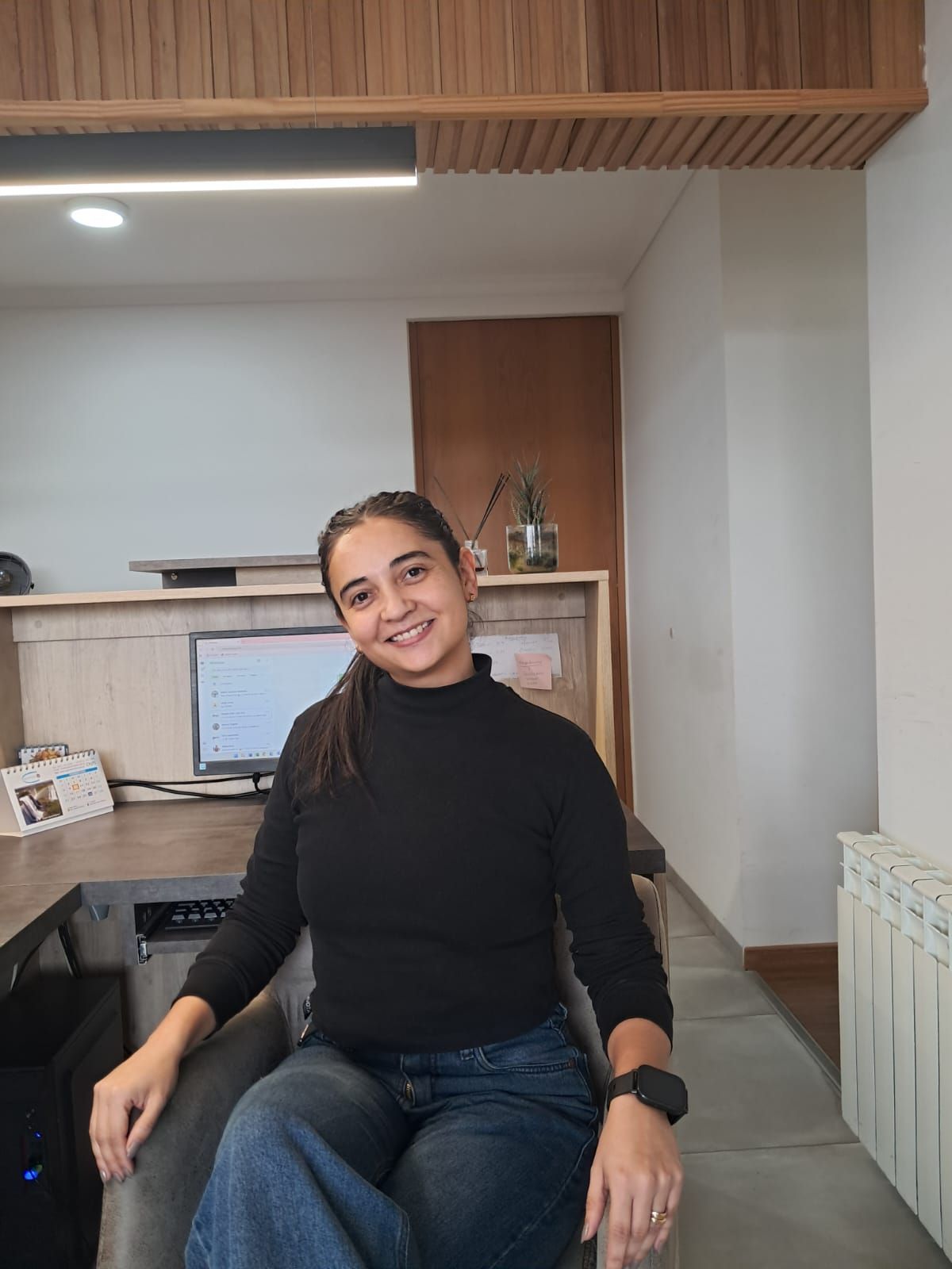 Mujer sentada en una silla negra, sonriendo en una oficina o sala de estar moderna con escritorio y estantes de madera.