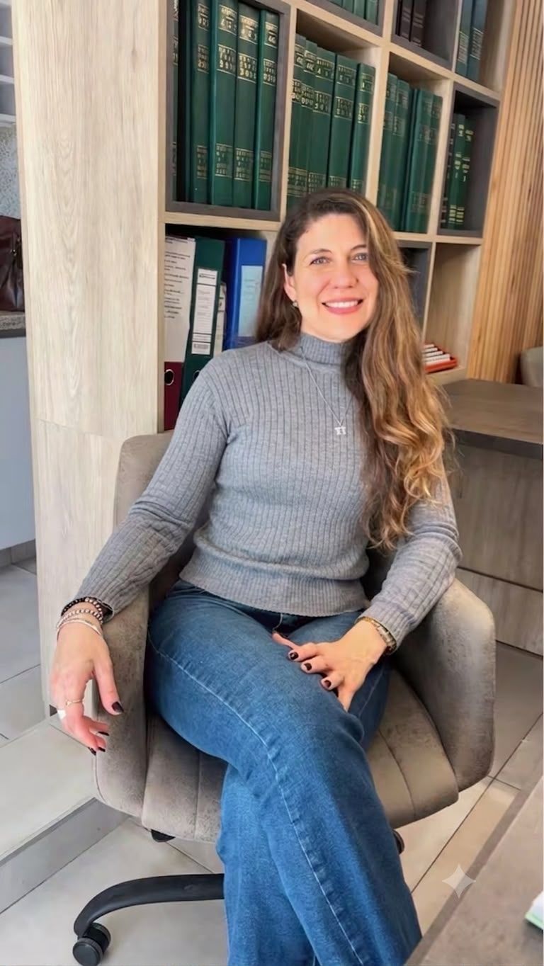 Mujer sentada en una silla gris junto a una estantería, vestida con un jersey gris y vaqueros, sonriendo en el despacho de su casa.