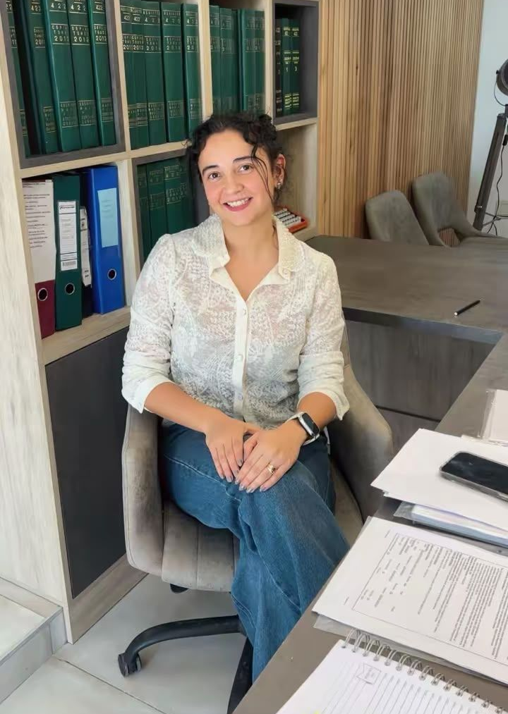 Mujer sentada en un escritorio de oficina, sonriendo junto a estantes llenos de carpetas y papeles.
