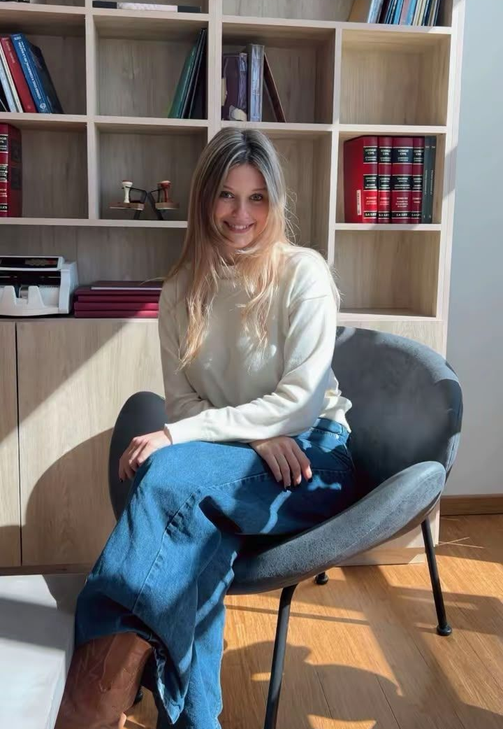 Mujer sentada en una silla gris junto a una estantería, con un suéter color crema y vaqueros azules, en una habitación iluminada por el sol.