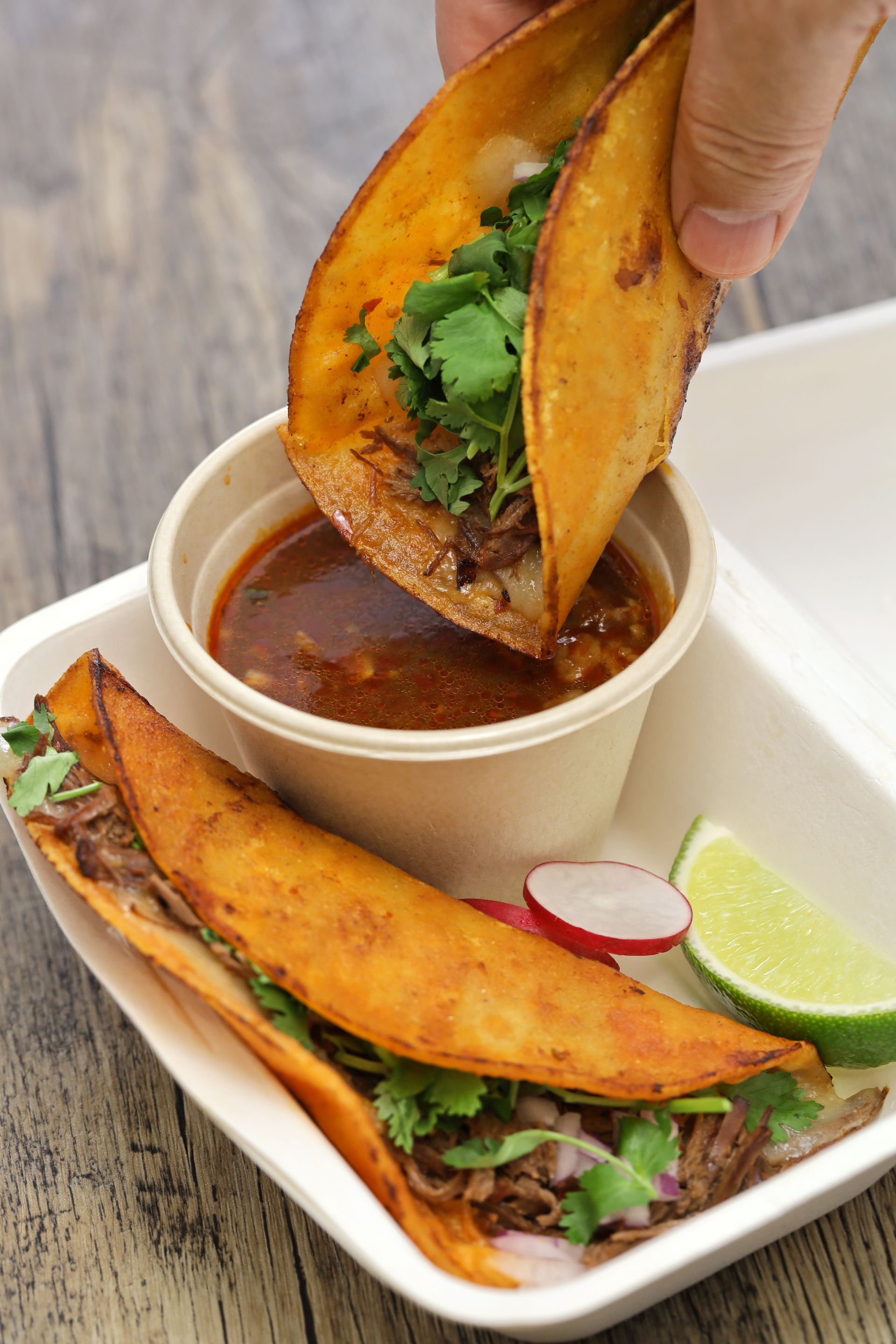 Birria tacos with consommé, cilantro, and radishes in a to-go container.