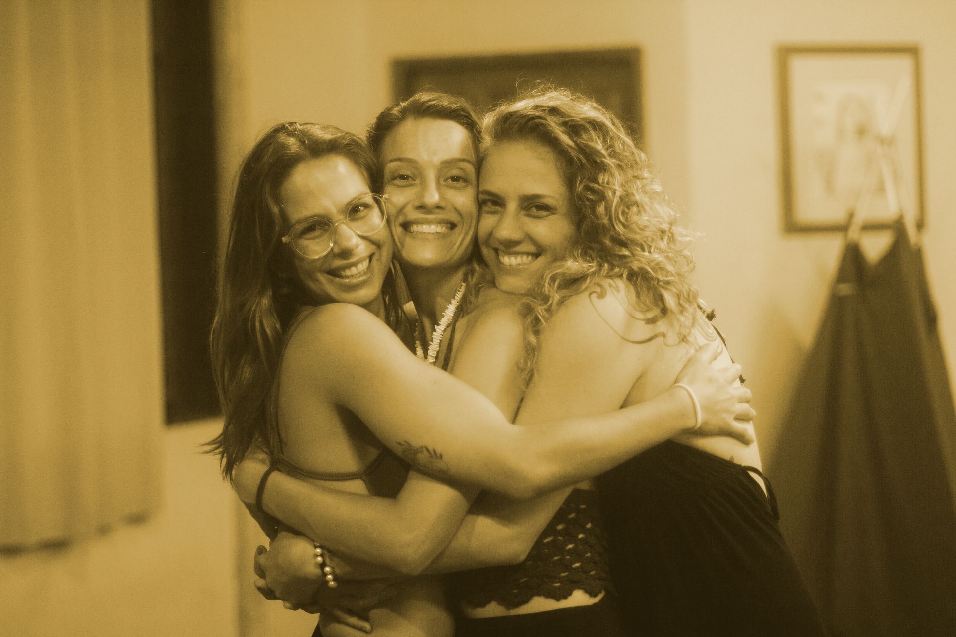 Três mulheres sorridentes se abraçando em uma sala com iluminação aconchegante, com um quadro e um casaco ao fundo.