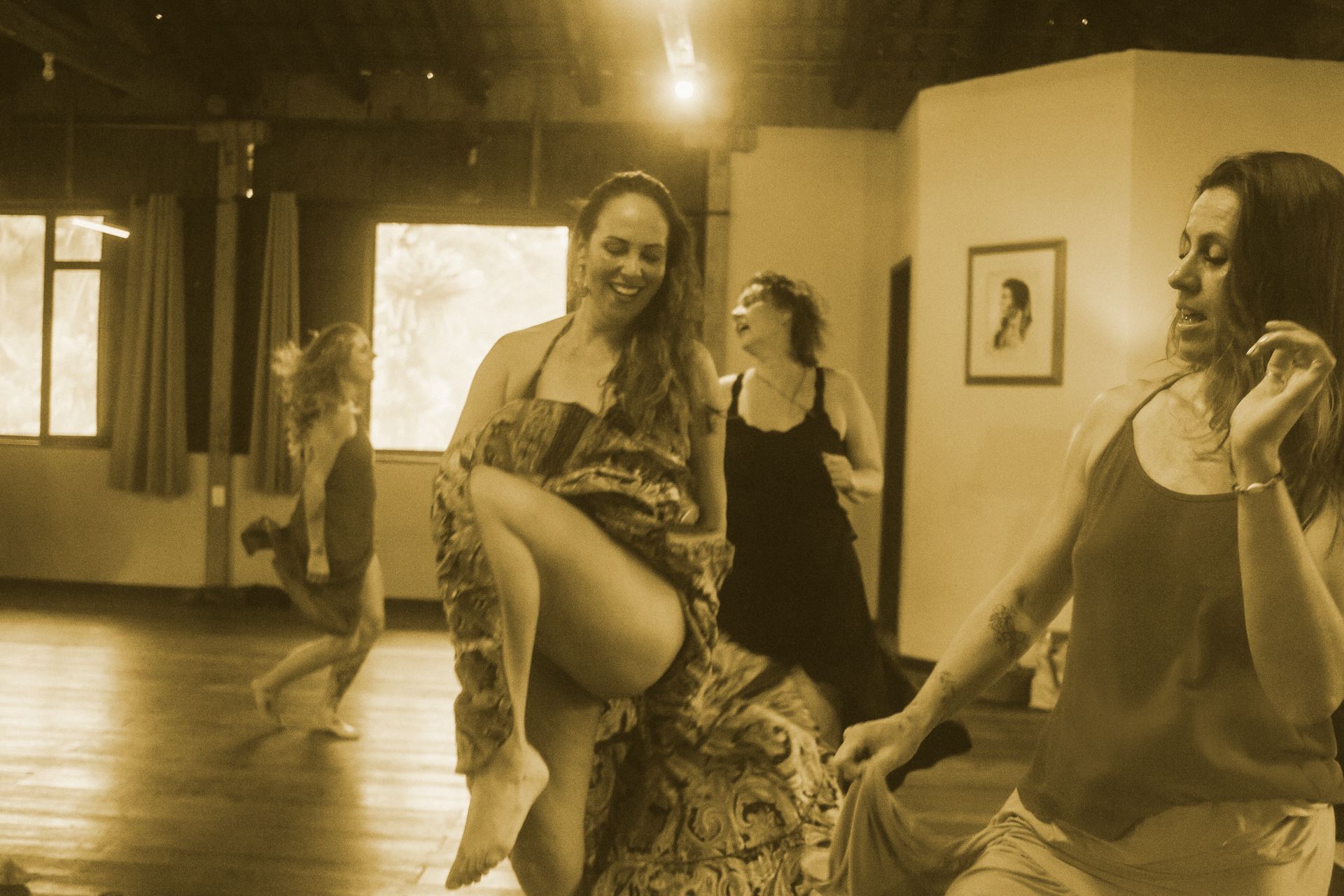 Mulheres dançando em um estúdio com iluminação aconchegante, uma delas sorrindo em primeiro plano com um tecido esvoaçante.