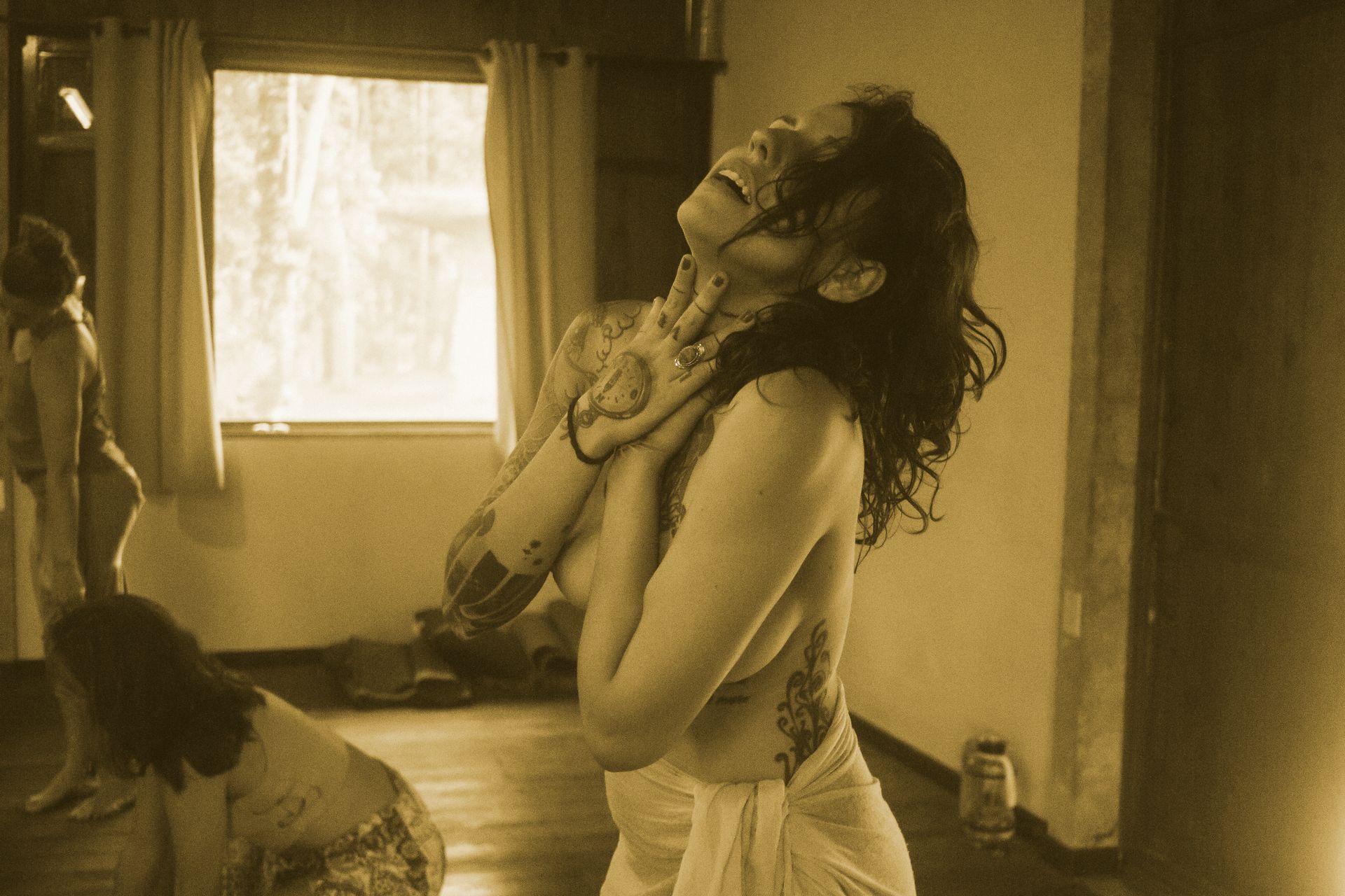 Mulher em um quarto em tons sépia, recostada com as mãos no peito perto de uma janela ensolarada.