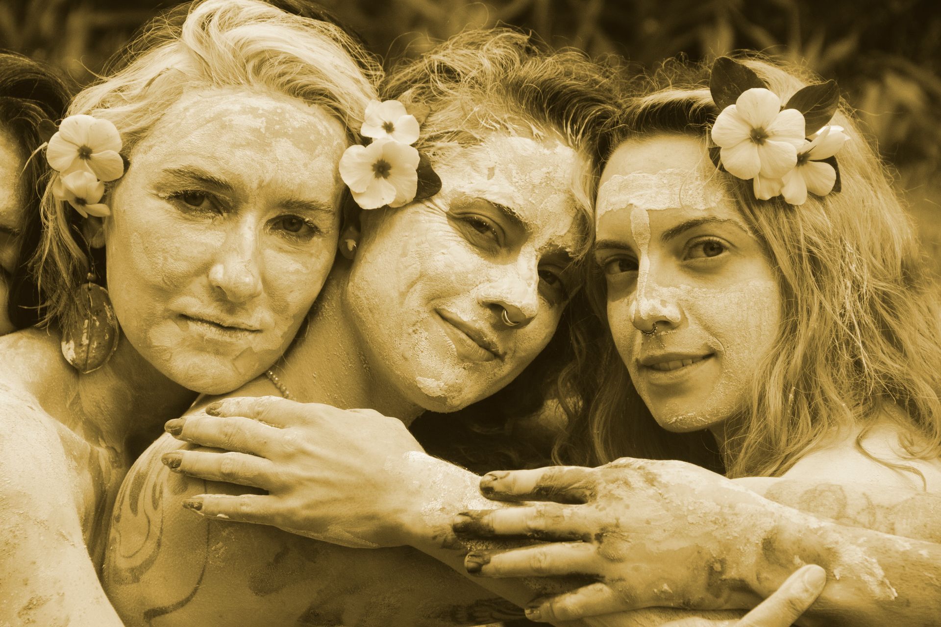 Três mulheres com coroas de flores e cabelos molhados se abraçam em um retrato em tons de sépia.