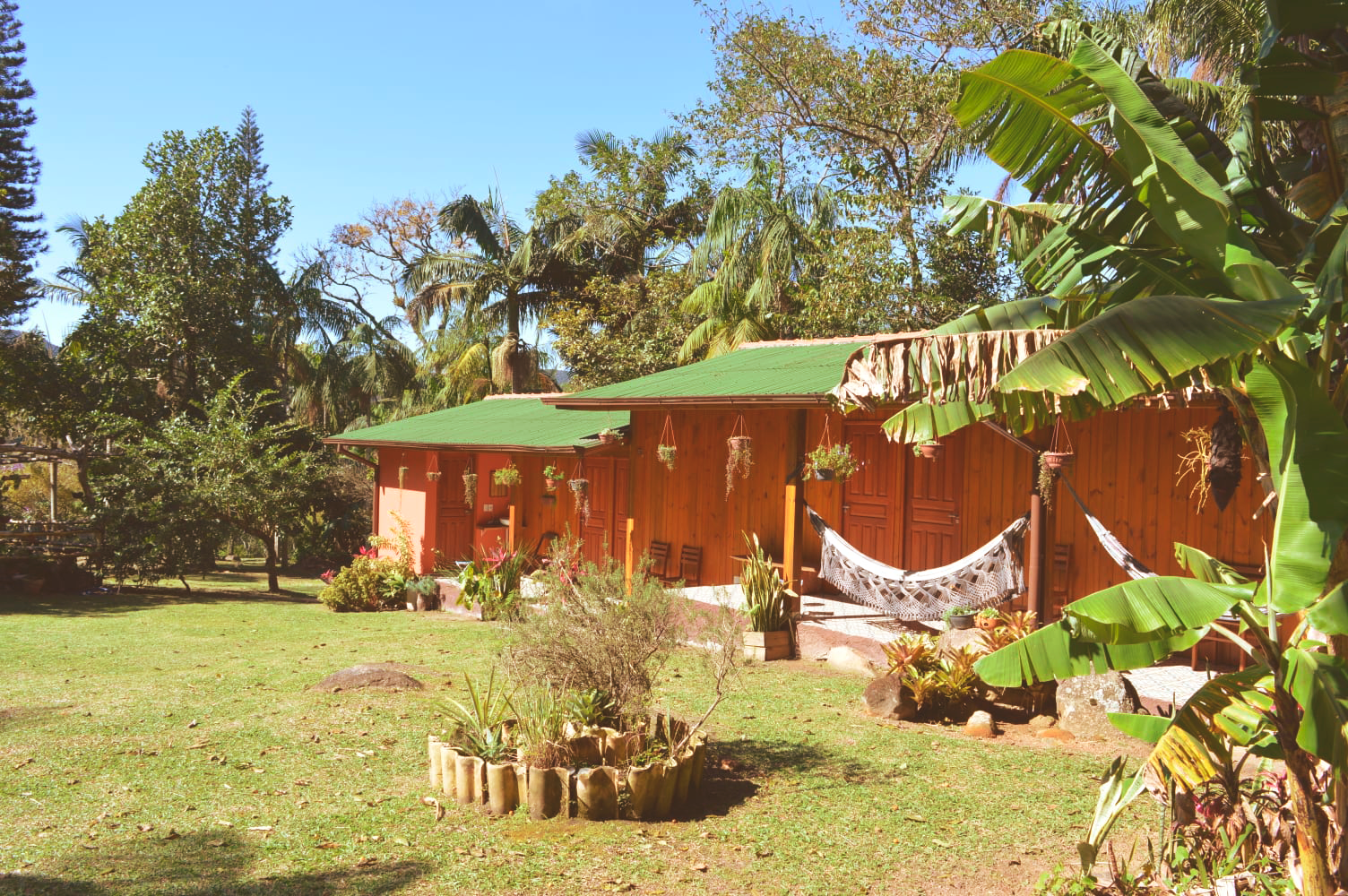 Casa laranja com telhado verde em um jardim ensolarado, rodeada por árvores e grama.