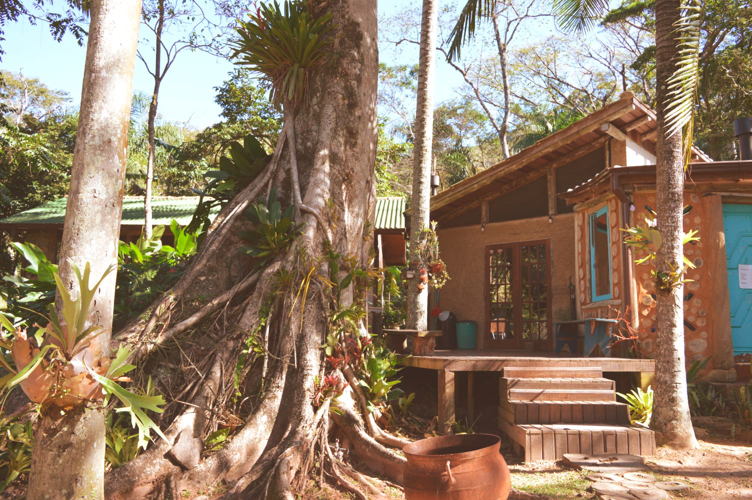 Cabana tropical com varanda, rodeada por palmeiras e vegetação exuberante, banhada por um sol radiante.
