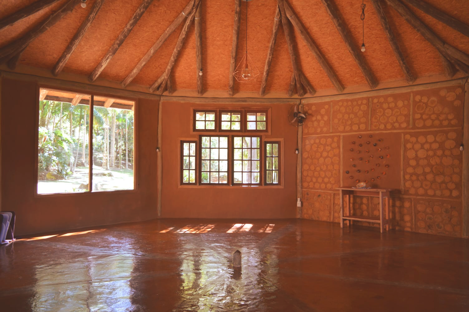 Sala circular vazia com piso de madeira, janelas e teto de palha, iluminada pela luz quente do sol.