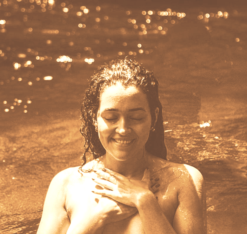 Pessoa sorrindo com os olhos fechados em água morna, mãos no peito, sob luz dourada.