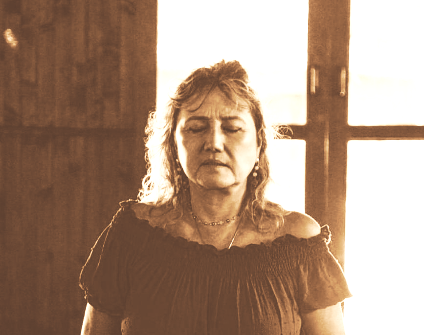 Mulher em pé junto a uma janela iluminada, olhos fechados, vestindo uma blusa escura de ombro caído.