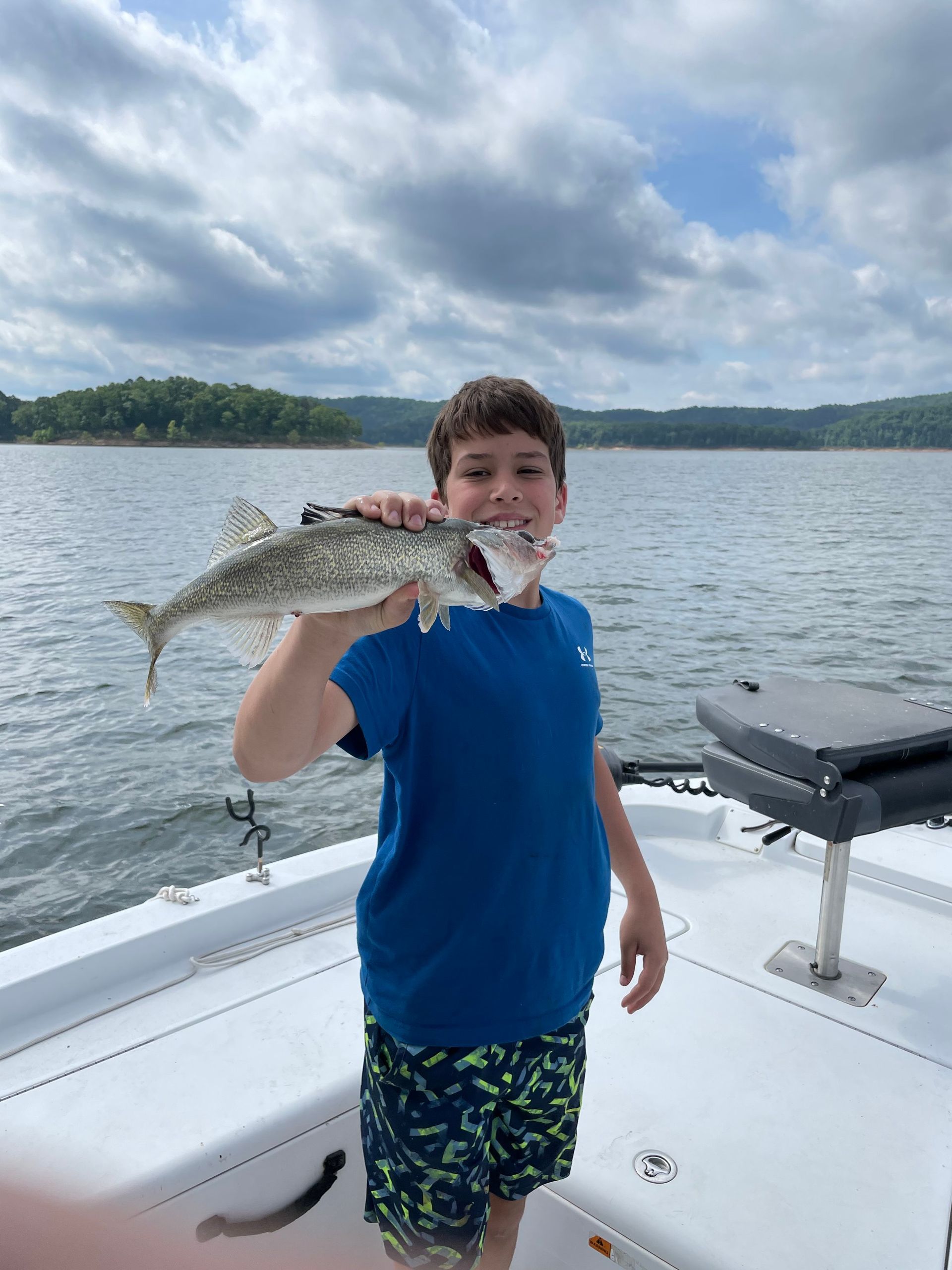 A young boy is standing on a boat holding a fish in his mouth.