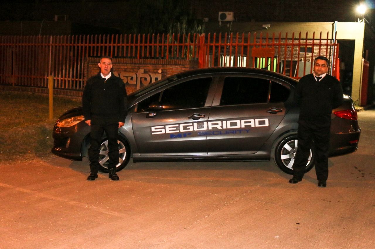 Dos guardias de seguridad están de pie junto a un coche gris por la noche; en el coche se lee 