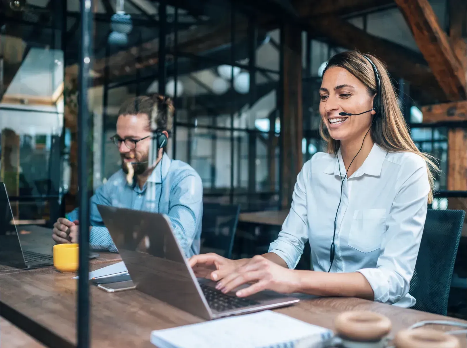 Two people wearing headsets, working on laptops at a wooden desk in an office.