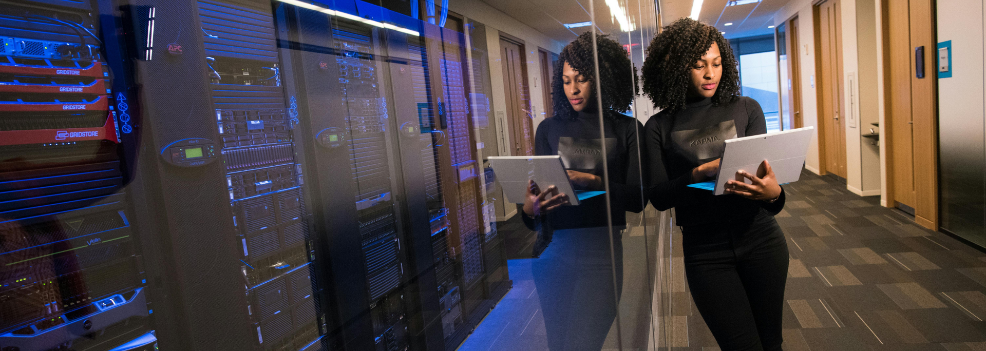 Woman in a dark outfit holding a tablet, standing in a hallway next to a glass wall with server racks.