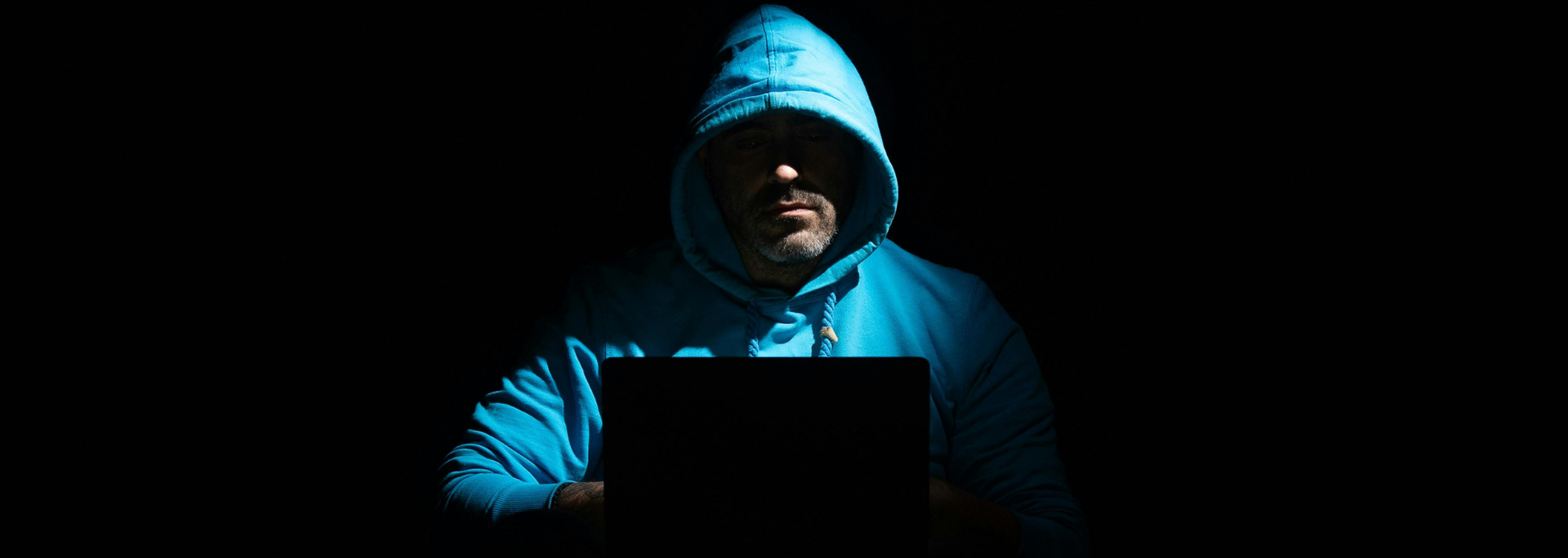 Picture of a hooded man sitting behind a laptop in a dark room. 