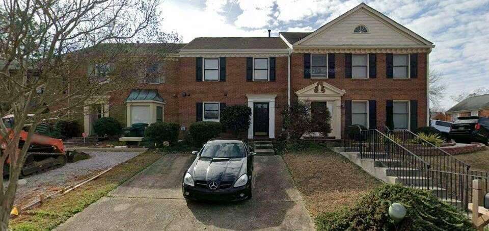Two-story brick townhouse with black car parked in driveway and bare trees in front yard.