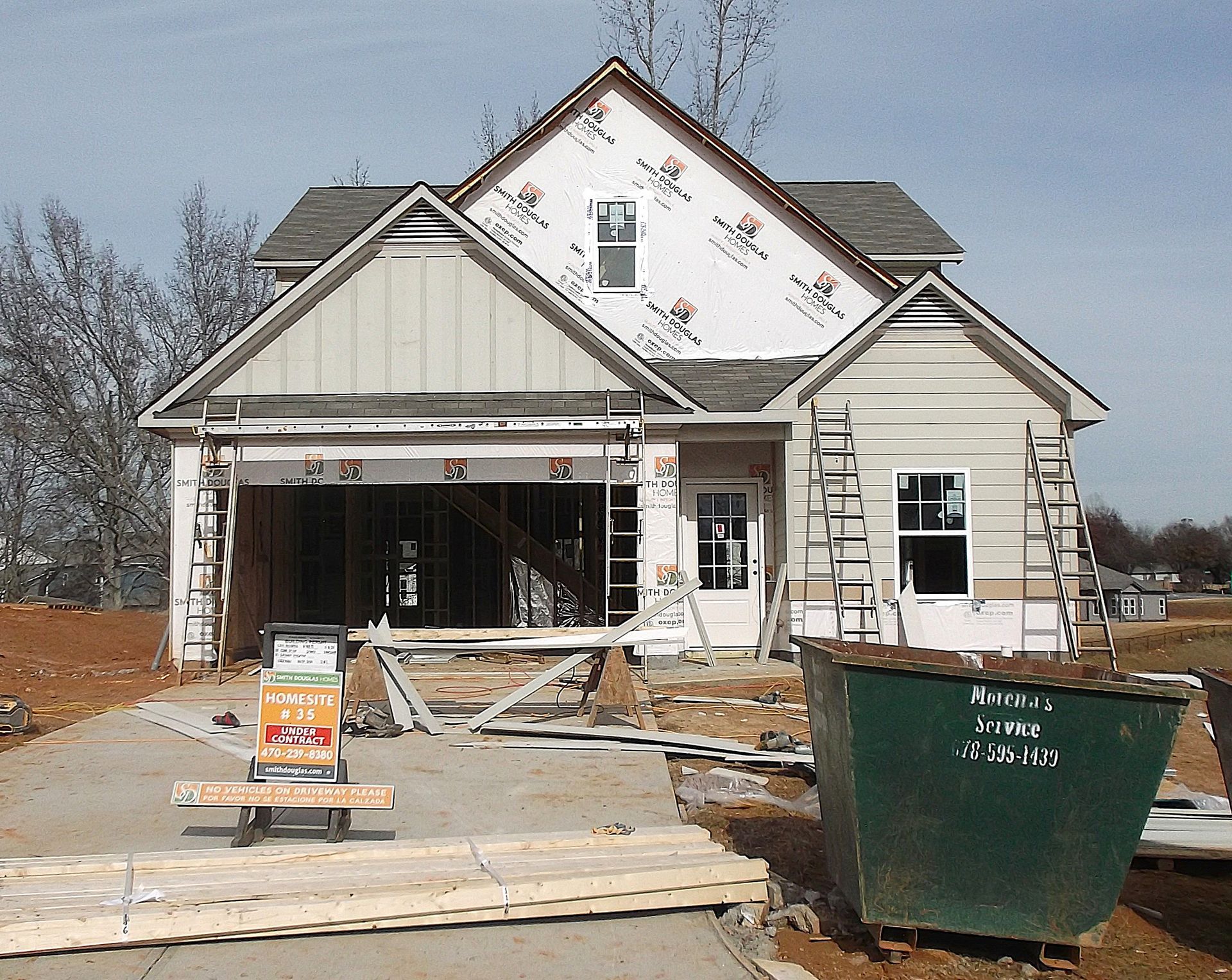 House under construction with open garage, framed porch, and a green dumpster in front.