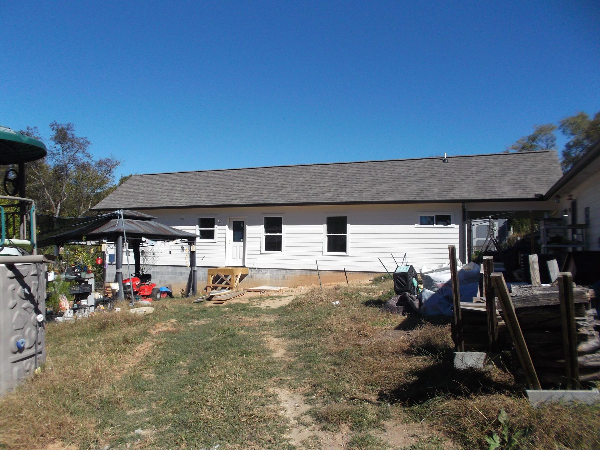 White single-story house with a gray roof and cluttered yard under a clear blue sky