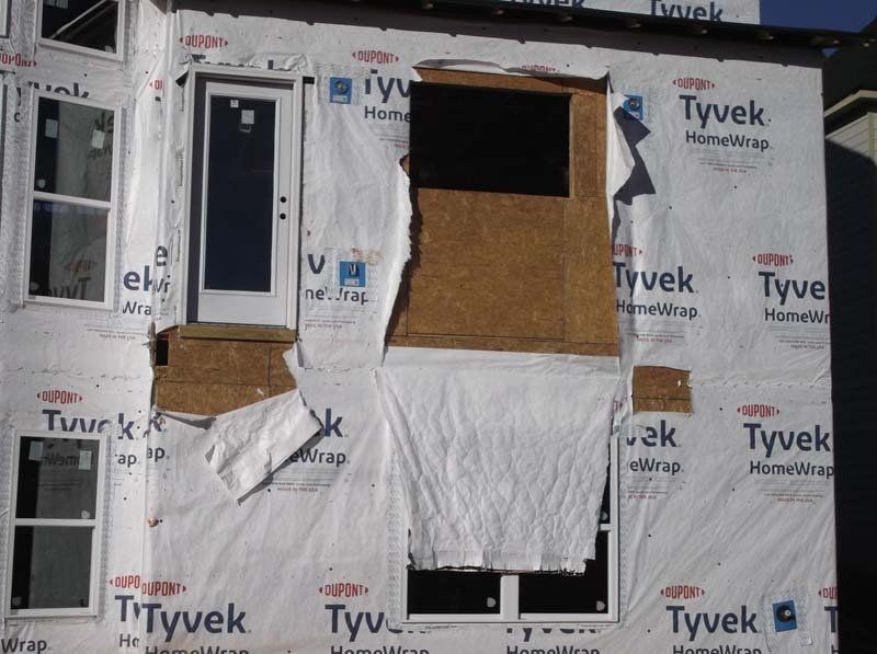 Partially covered house exterior with Tyvek wrap, boarded windows, and a window opening