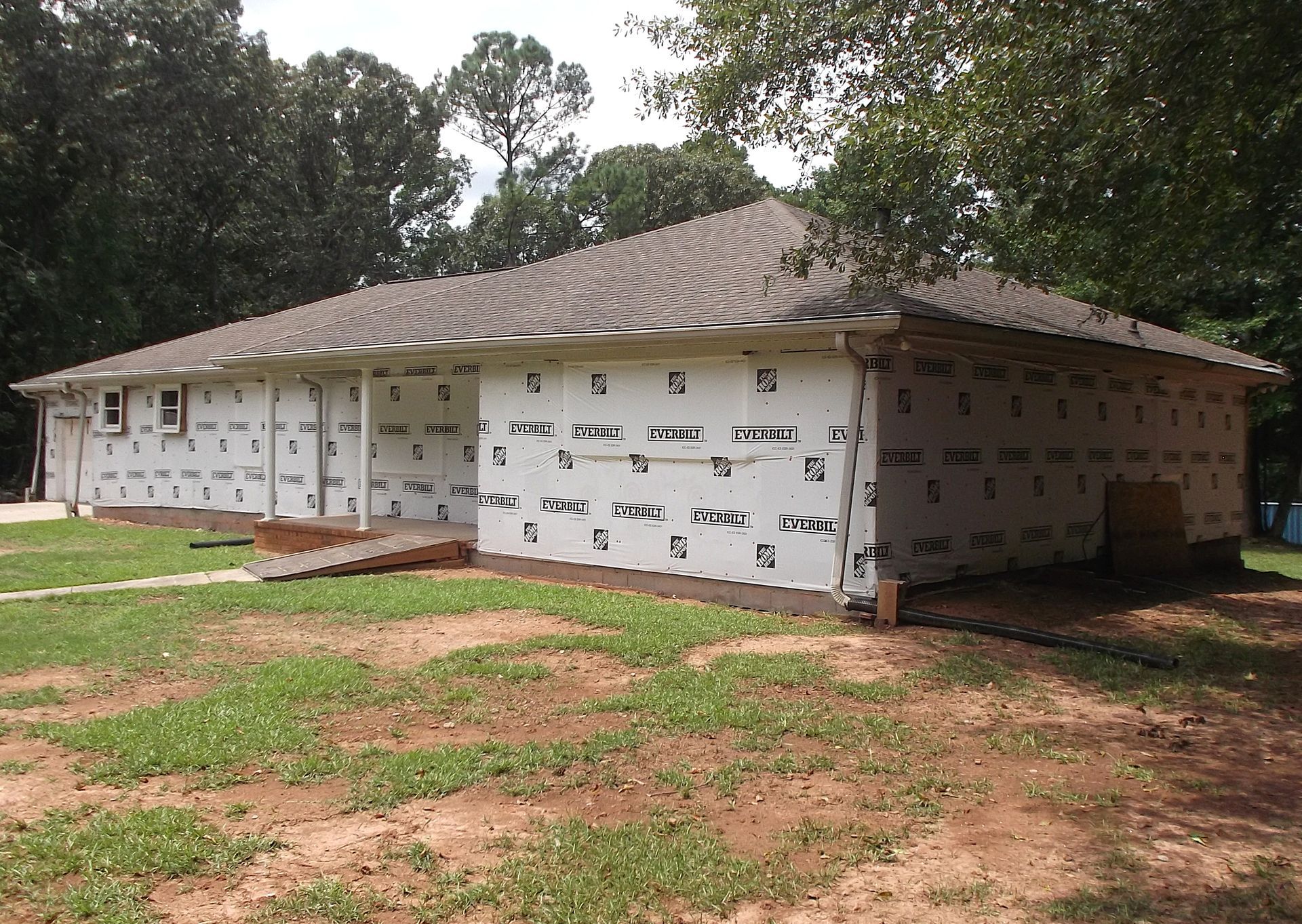 Partially built white house with a dark shingled roof in a wooded dirt lot