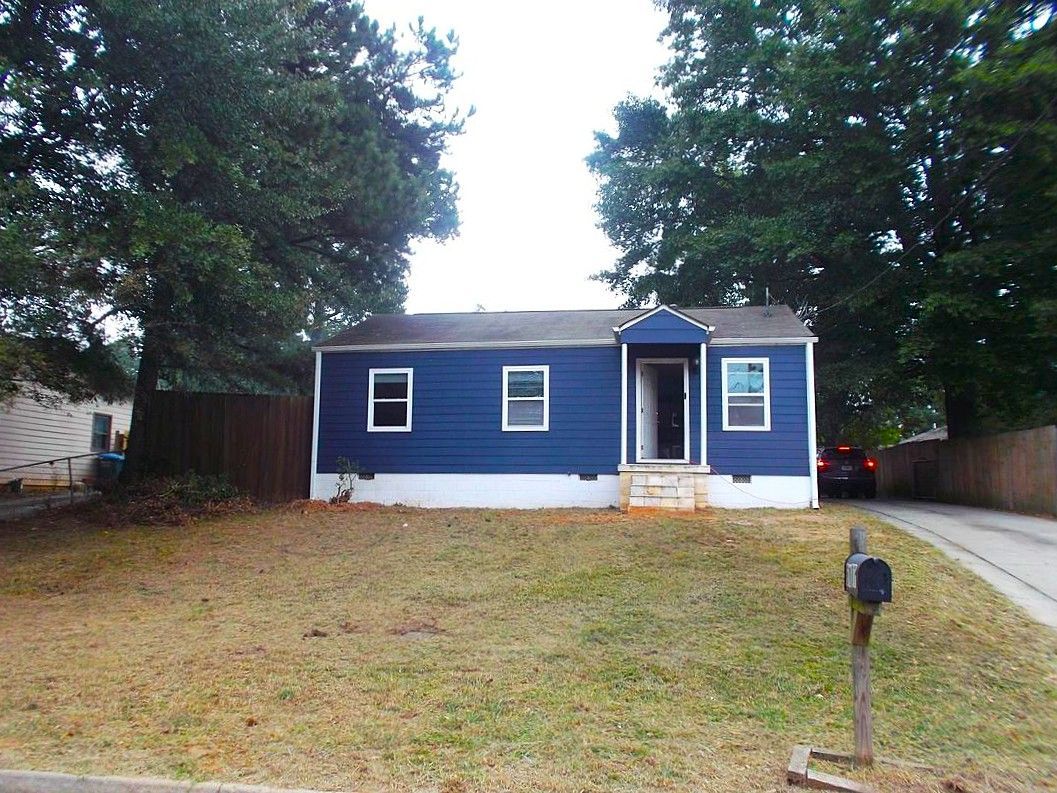 Small blue house with white trim, front porch, and driveway in a grassy yard with tall trees.