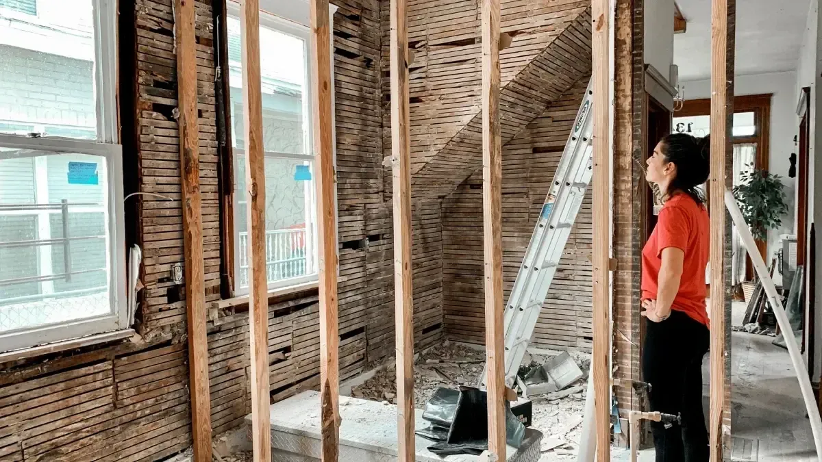 Interior renovation with exposed wall studs, debris, ladders, and a person in a red shirt inspecting the room