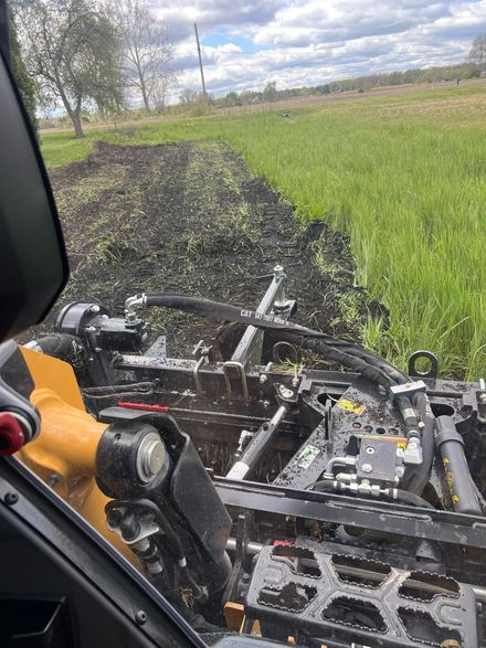 View from a farm vehicle cab over a black tilled field beside green grass under cloudy sky