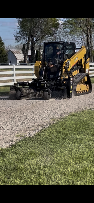 Yellow backhoe pushing gravel along a driveway beside a white fence and green lawn.