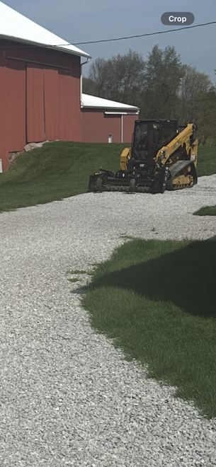 Yellow backhoe on a gravel path beside a red barn and grassy hill under a cloudy sky