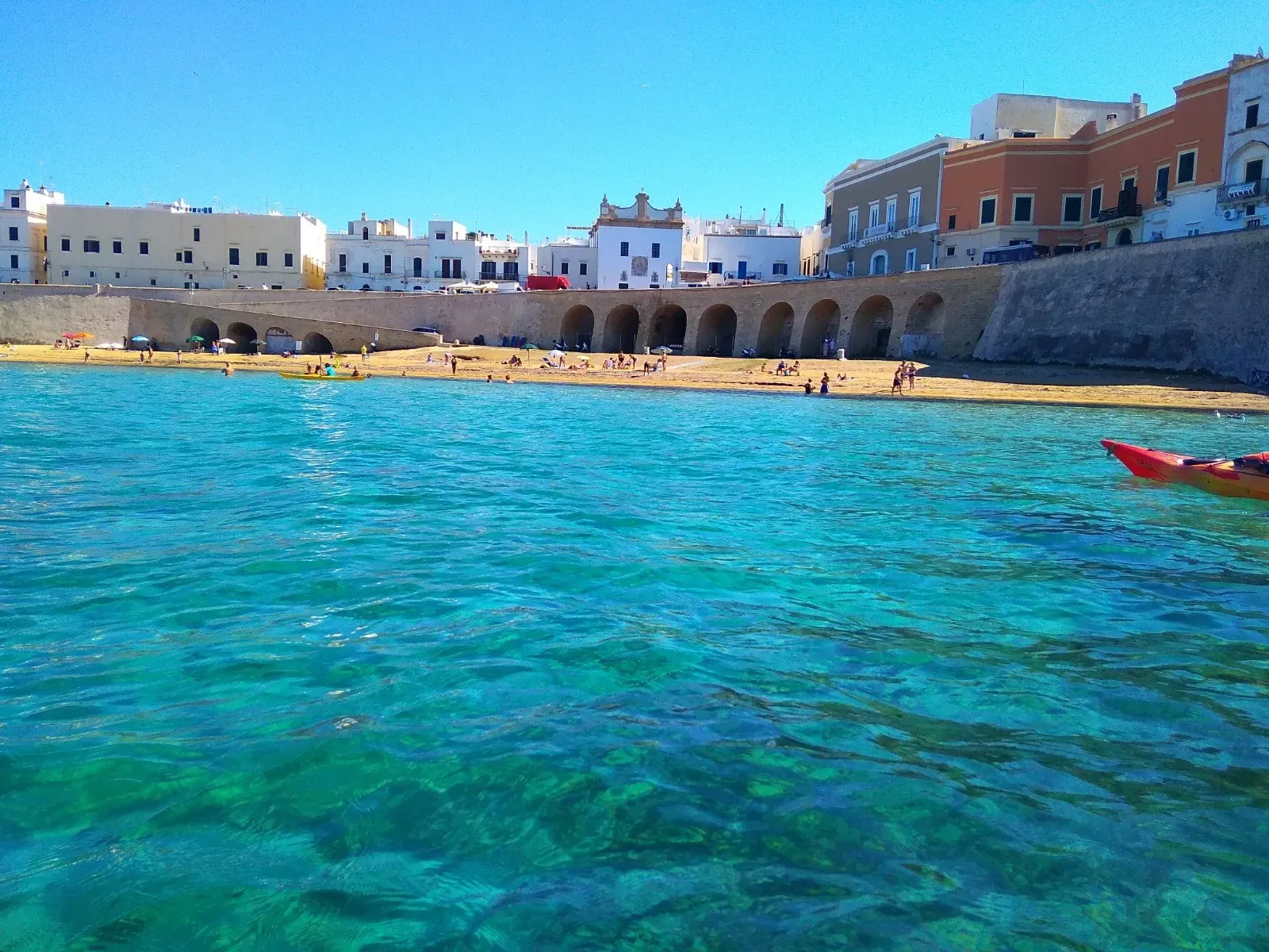 Mare turchese accanto a un lungomare di pietra con edifici bianchi e una barca rossa sotto un cielo azzurro limpido.