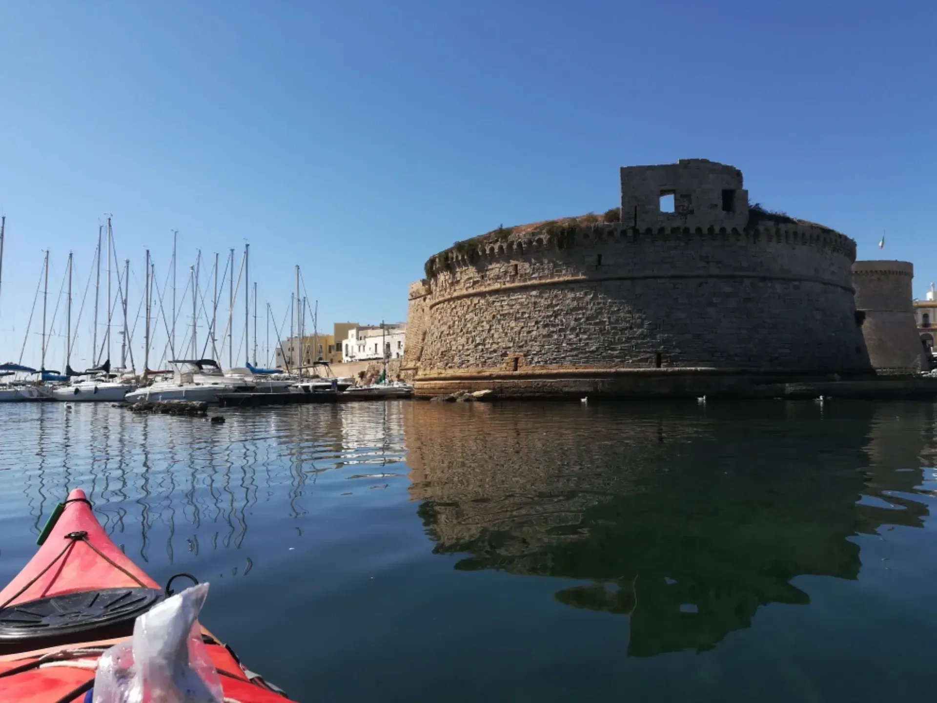 Forte portuale in pietra accanto ad acque calme con barche a vela e un kayak rosso in primo piano.