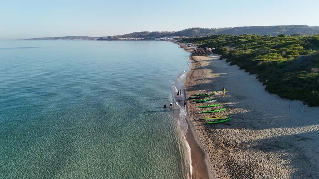 Veduta aerea di una tranquilla spiaggia con acqua cristallina, sabbia di ciottoli e alberi lungo la costa.