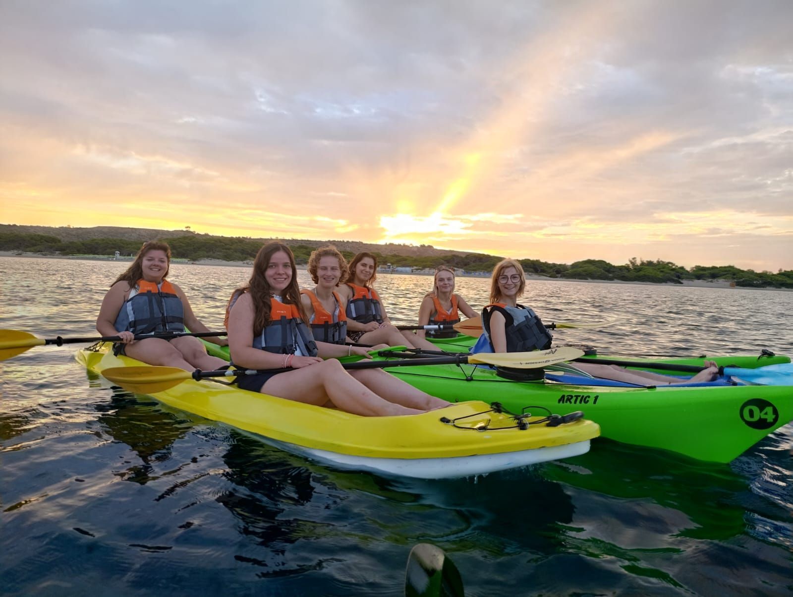 Cinque persone in kayak su un lago tranquillo al tramonto, con kayak colorati e un cielo luminoso.