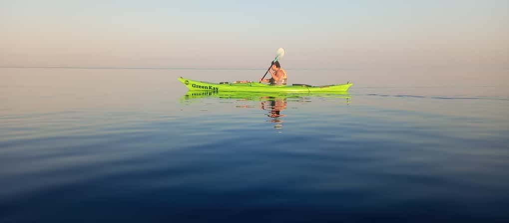 Una persona in un kayak verde brillante su acque calme all'alba, riflessa sulla superficie liscia come uno specchio.
