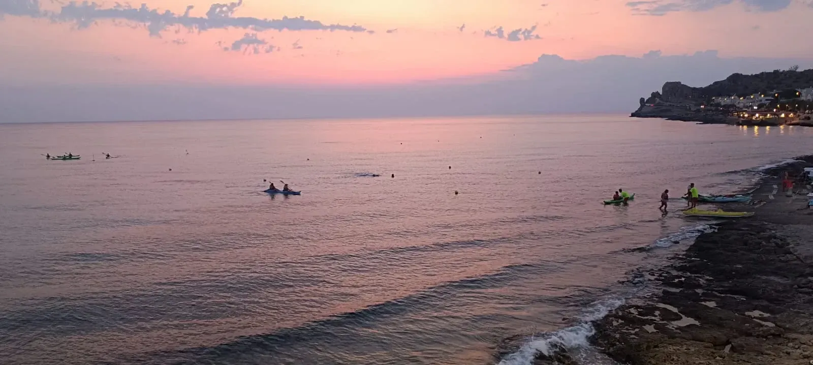 Tramonto rosa su un oceano calmo, con piccole barche vicino a una costa rocciosa e colline in lontananza.