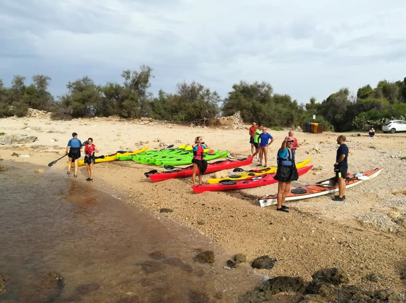 Persone con kayak sulla riva rocciosa di un fiume, pronte a varare imbarcazioni colorate in acque poco profonde.