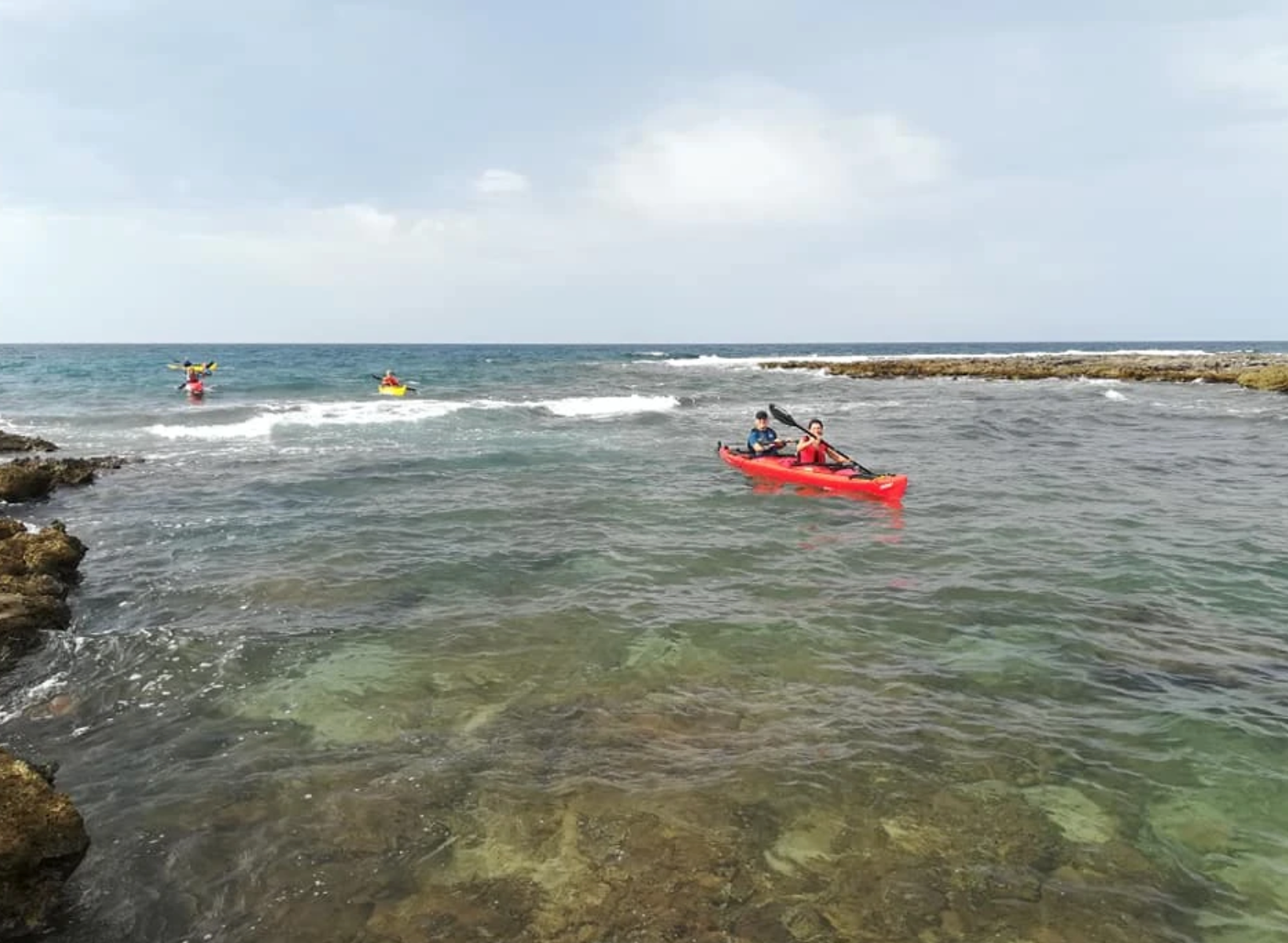 Persone in kayak in un mare poco profondo e roccioso vicino a una diga frangiflutti sotto un cielo nuvoloso.