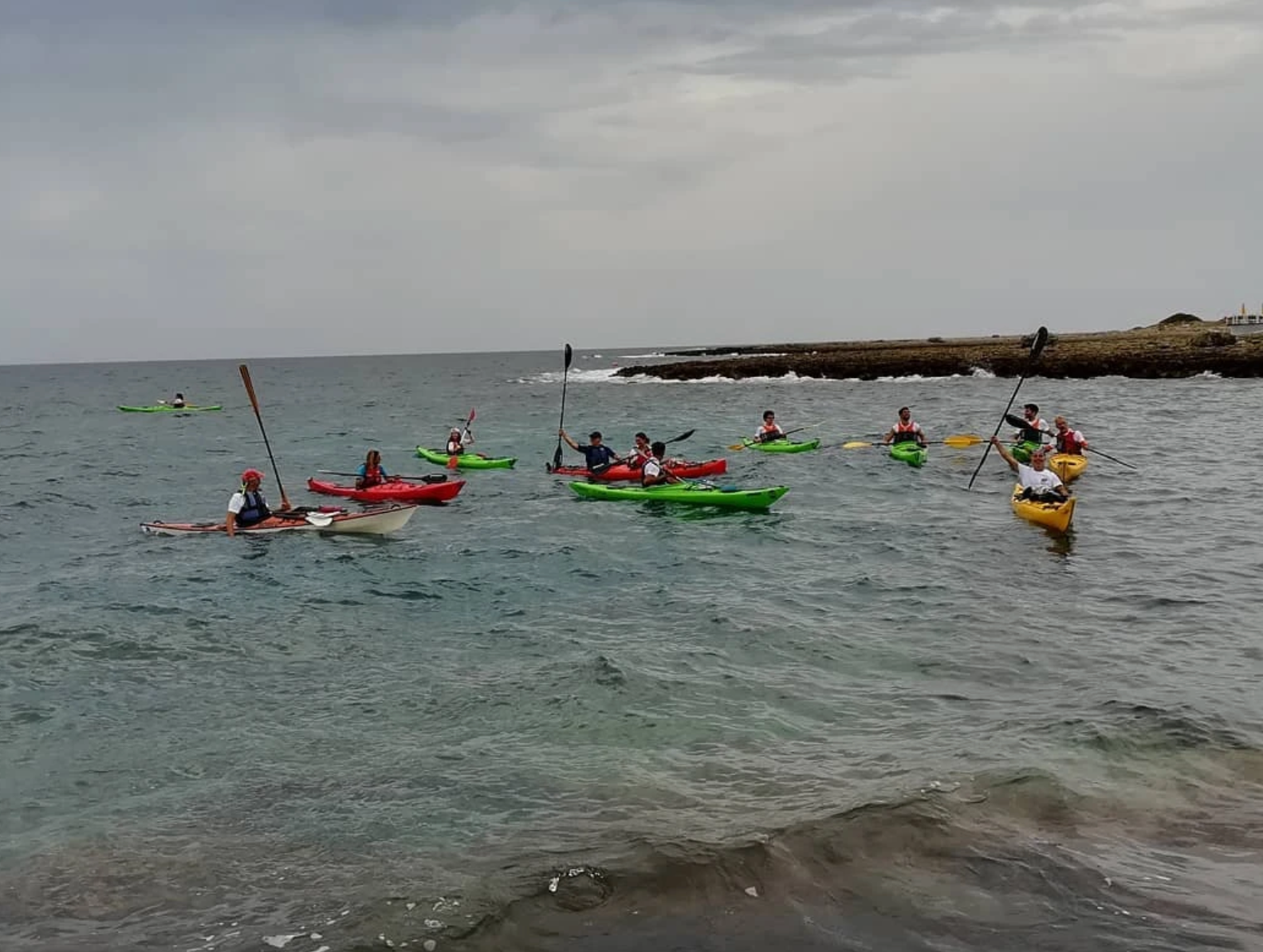 Kayakisti che remano in acque agitate vicino a una costa rocciosa sotto un cielo nuvoloso.