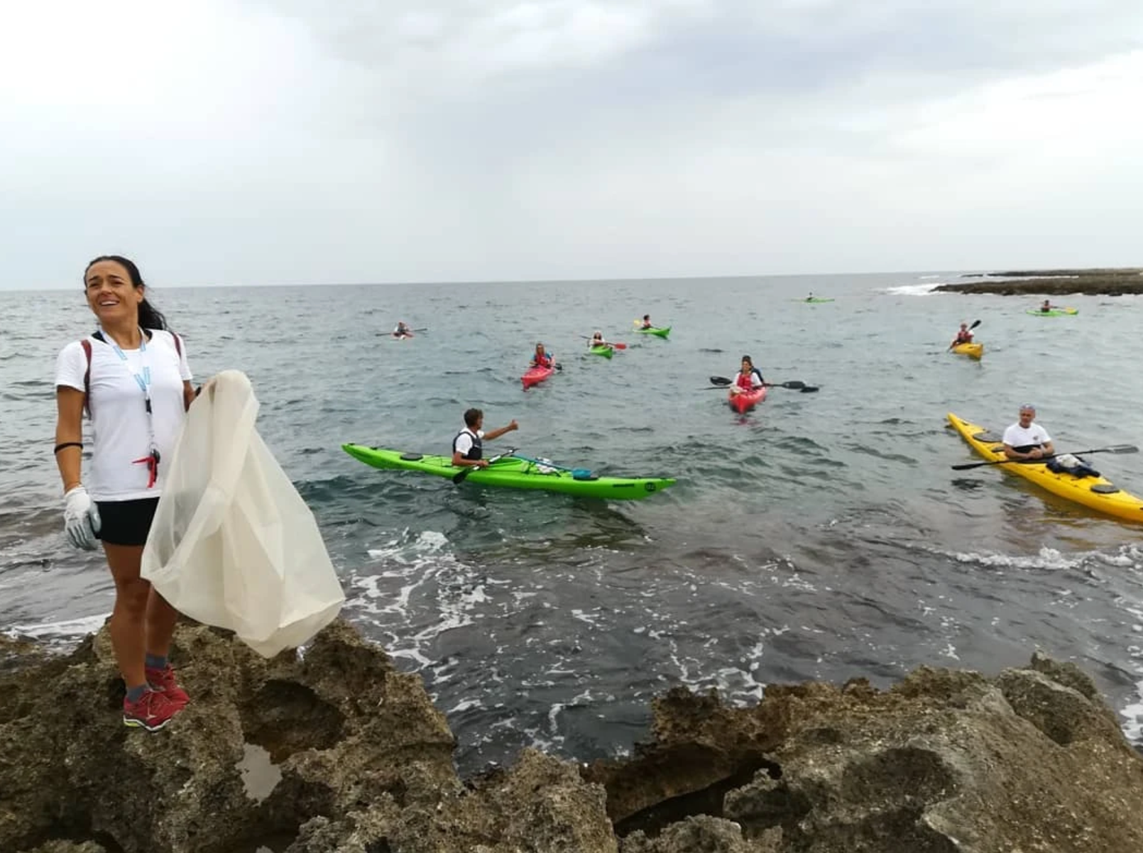 Una donna su una riva rocciosa vicino a dei kayakisti su kayak colorati in acque calme sotto un cielo nuvoloso.