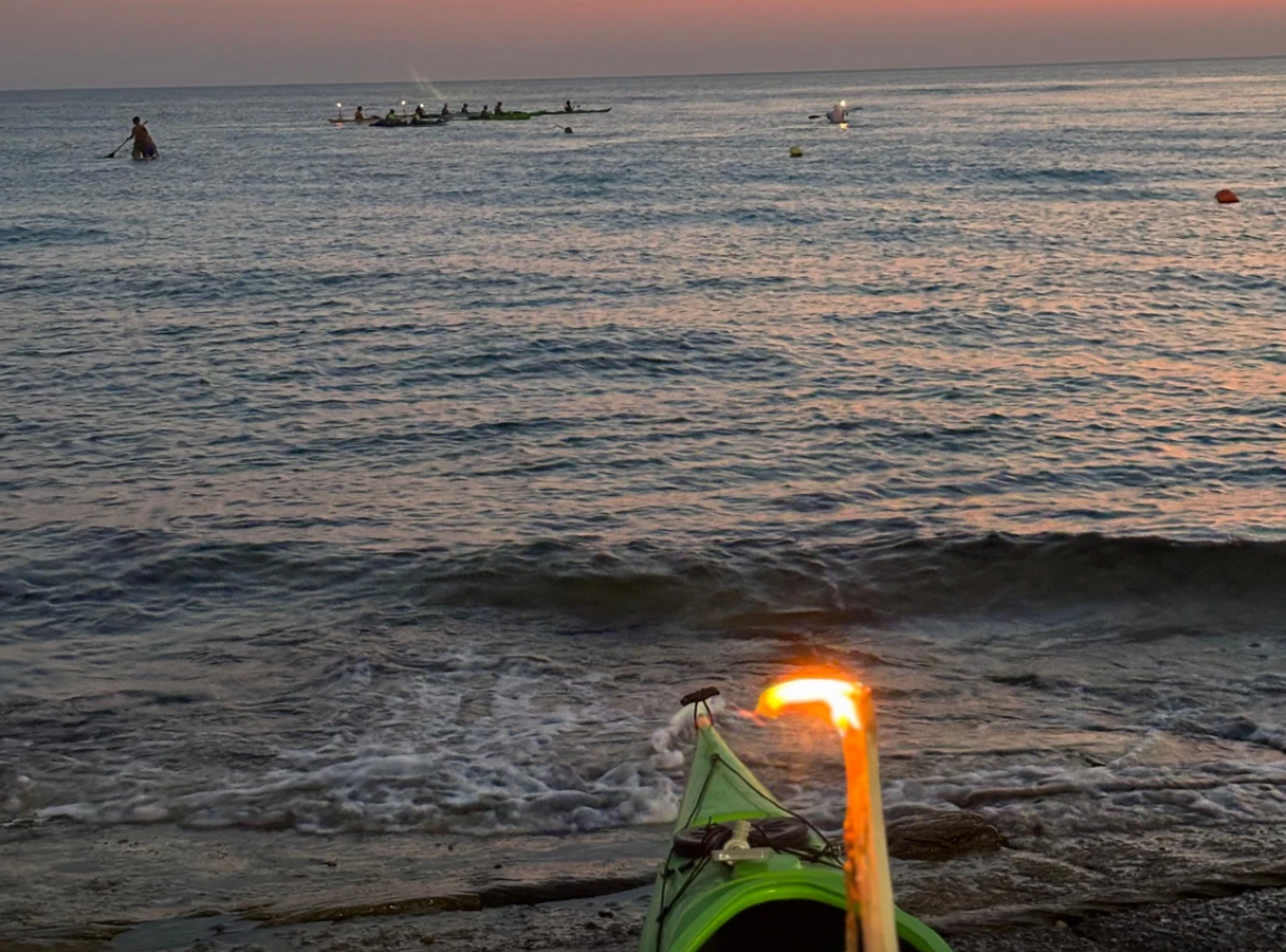 Prua di kayak illuminata, rivolta verso un oceano calmo al tramonto, con surfisti in lontananza all'orizzonte.
