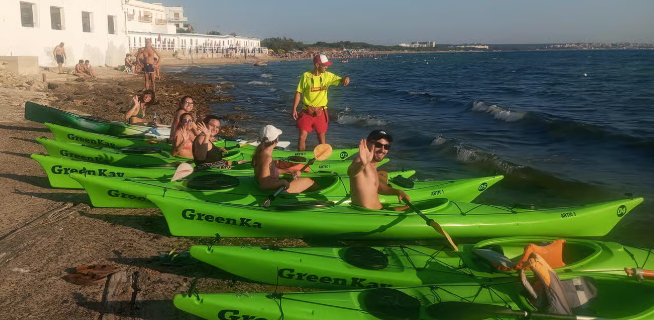 Gruppo di persone su kayak verde brillante in riva al mare, con una persona in piedi sulla spiaggia alle loro spalle.