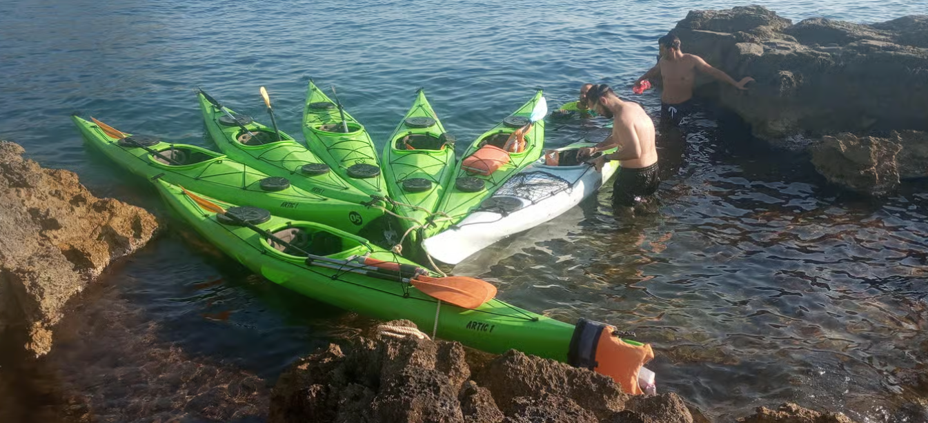Una persona in un kayak verde con attrezzatura a forma di foglia, galleggia vicino a delle rocce su acque calme.