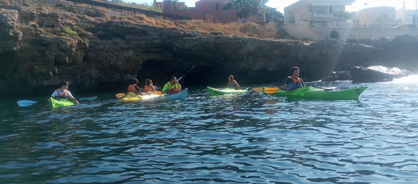 Kayakisti che remano sulle acque azzurre vicino a una costa rocciosa, con alcune case sullo sfondo.