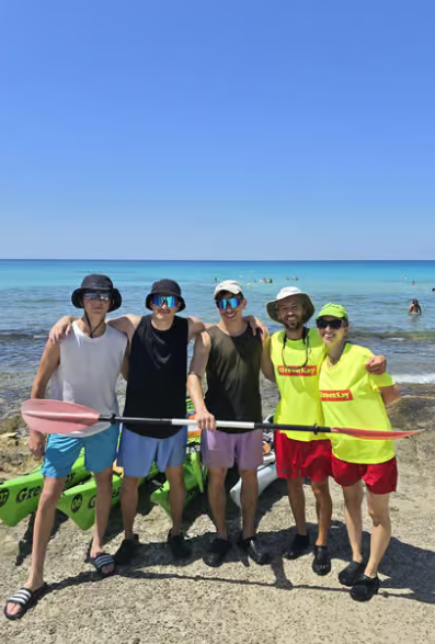 Cinque persone in posa con un nastro rosso su una spiaggia assolata, con l'oceano e il cielo azzurro sullo sfondo.