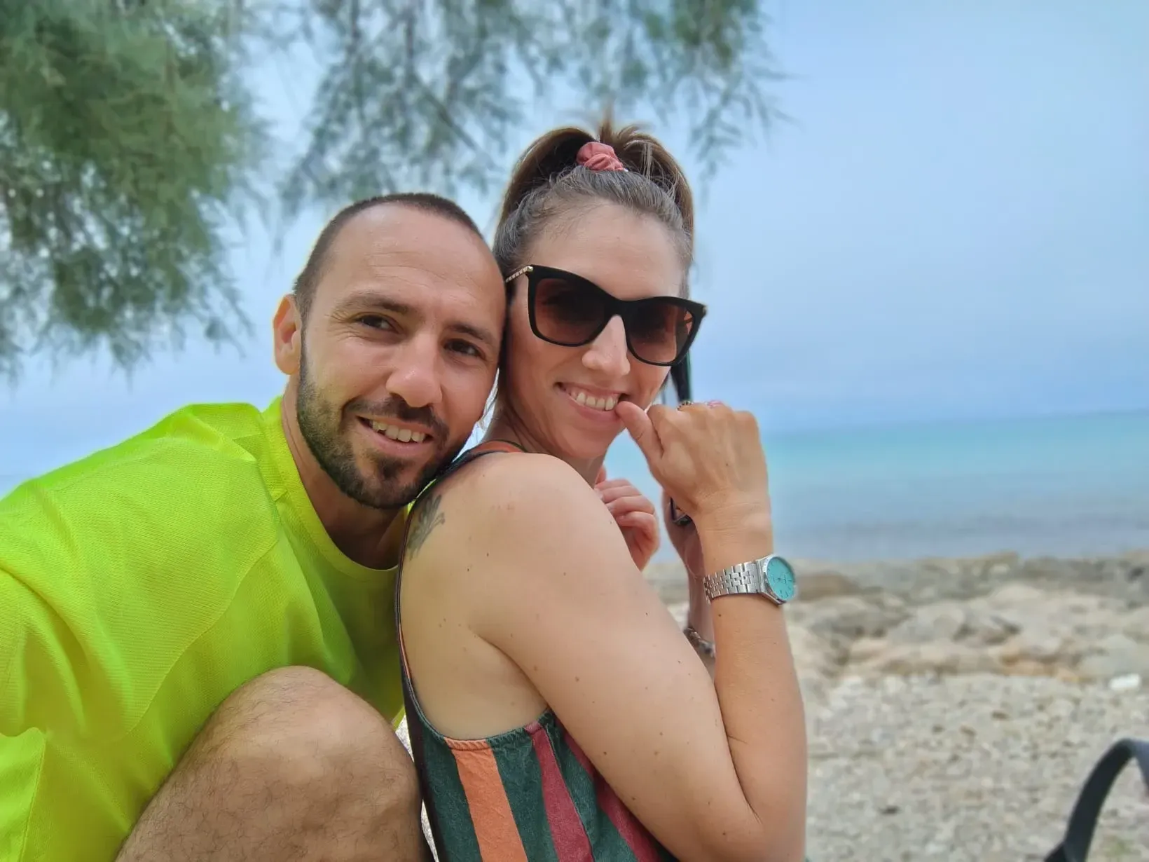 Due persone sorridenti su una spiaggia, una con una maglietta verde fluo e l'altra con gli occhiali da sole.