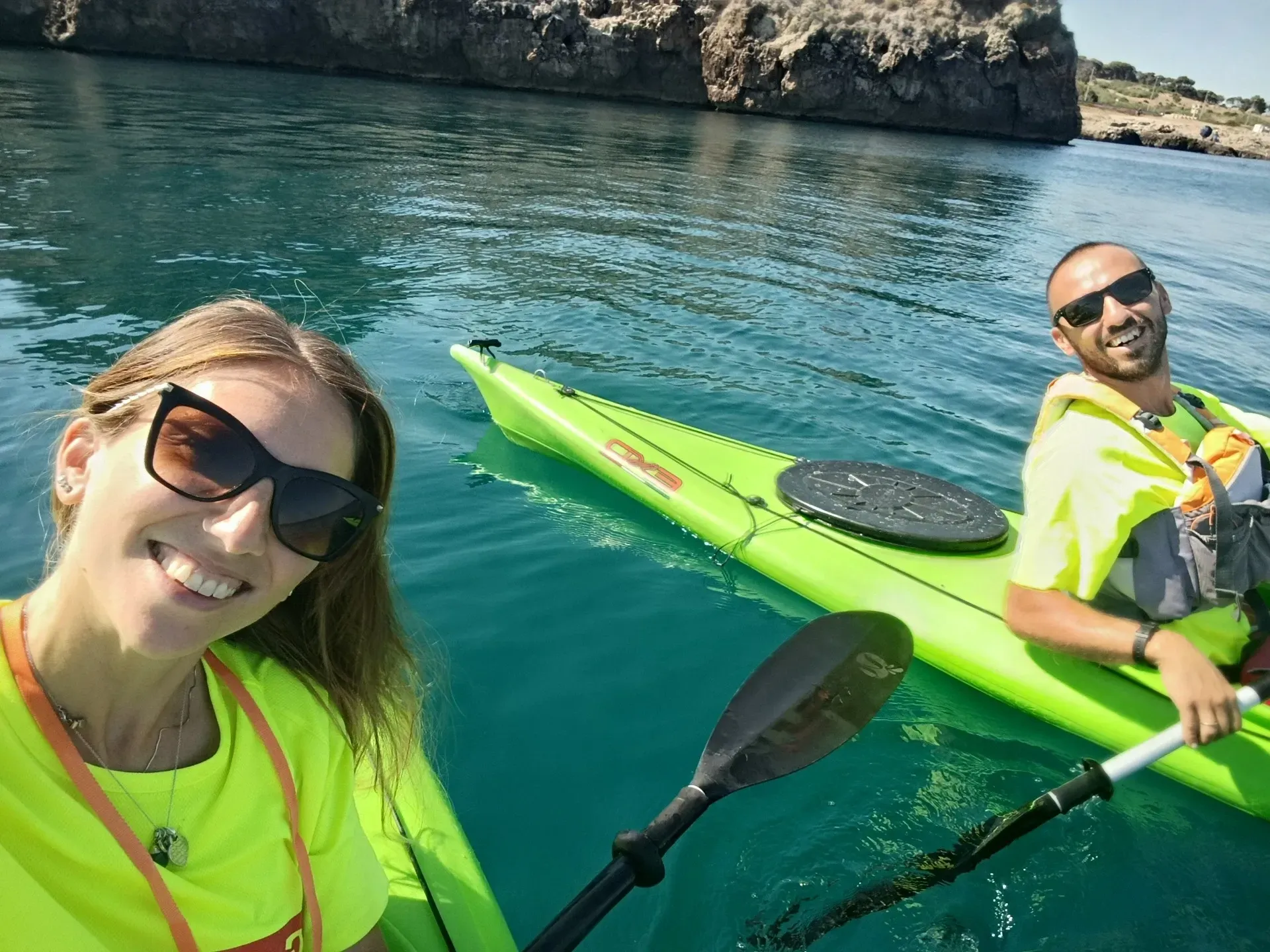 Due kayakisti su kayak verde brillante sorridono sulle acque cristalline vicino a scogliere rocciose.