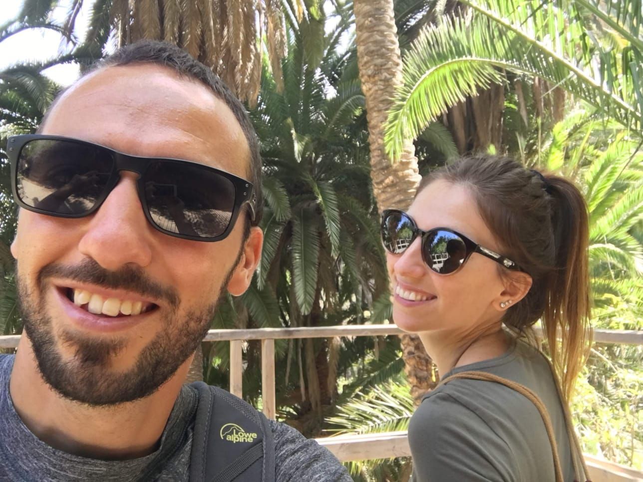Due persone sorridenti su una passerella tropicale, con palme sullo sfondo.