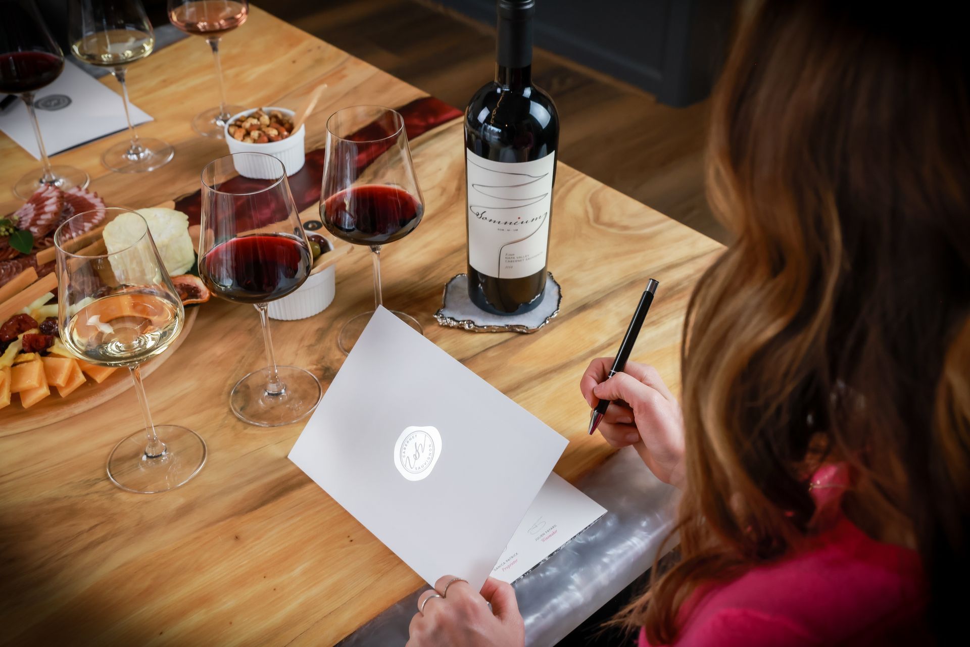 Woman writing at a table with wine glasses, charcuterie, and a wine bottle.