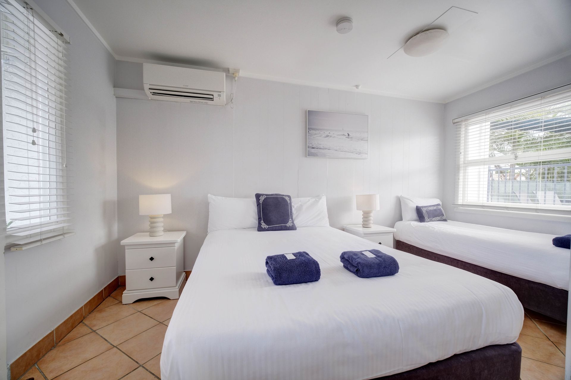 Bedroom with white walls, a double bed, and a single bed. Towels on the bed, two windows.