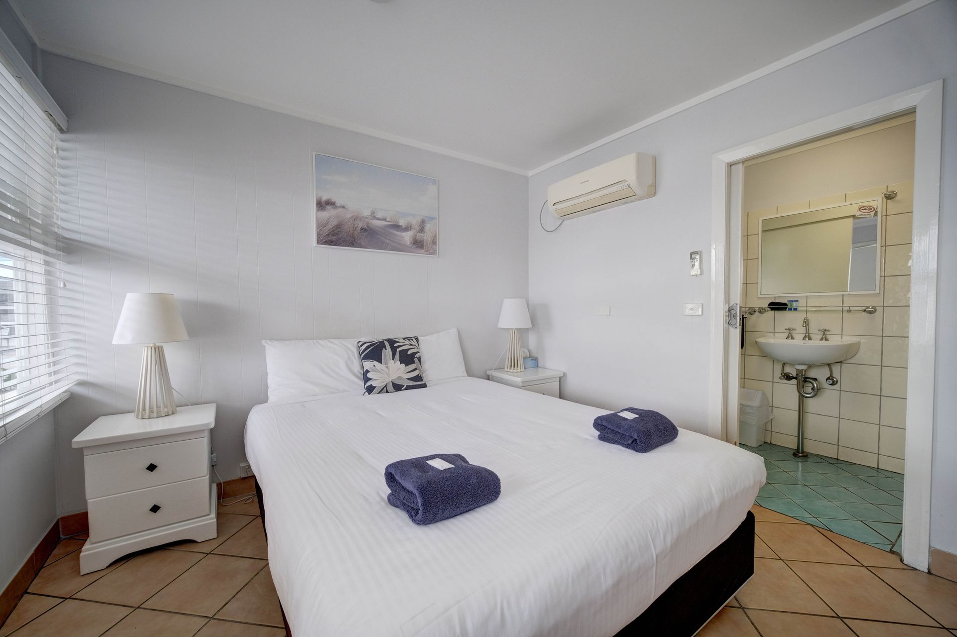 Bedroom with bed, bedside tables, window with blinds, and doorway to bathroom.